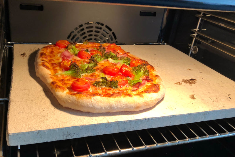 Pizzastein im Backofen mit Tomaten, Broccoli und Schinken belegt
